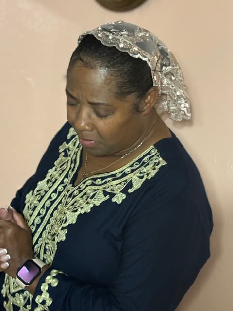 Woman praying with closed eyes, wearing a navy blue embroidered dress and lace veil.