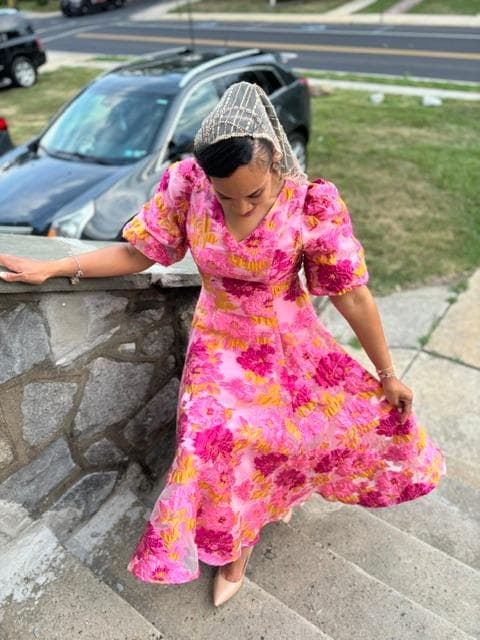 Woman in a vibrant pink floral puff-sleeve dress and beaded headpiece on stone steps.